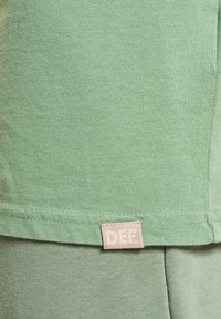 Lichtgroen katoenen t-shirt met een rechte onderrand en een klein beige label met "DEF" gestikt op de linkeronderkant. Zachte textuur.