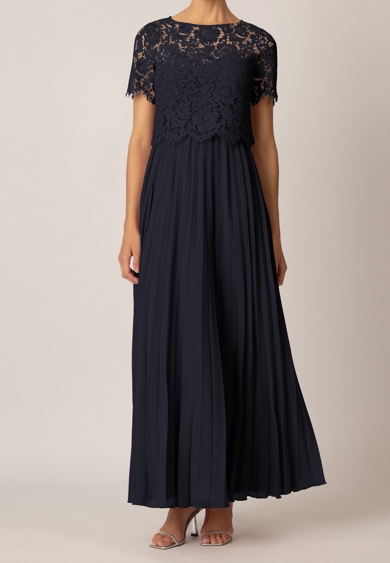 Femme portant une robe maxi bleu marine avec un haut en dentelle à manches courtes et une jupe plissée, associée à des talons ouverts argentés.