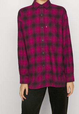 Chemise à manches longues avec un motif à carreaux rouge et noir, poche avant, portée avec un pantalon noir.
