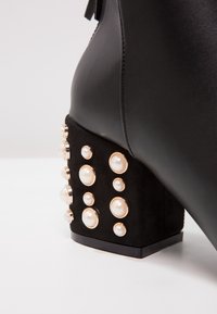 Schwarze Knöchelstiefelette mit einem strukturierten Wildleder-Blockabsatz, verziert mit goldenen und cremefarbenen Perlenakzenten, und einer glatten Lederoberfläche.
