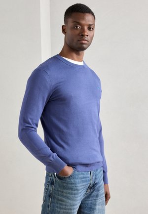Hellblauer Strickpullover mit langen Ärmeln, rundem Ausschnitt und gerippten Bündchen. Verfügt über ein kleines Logo auf der Brust. Kombiniert mit hellblauen Jeans.