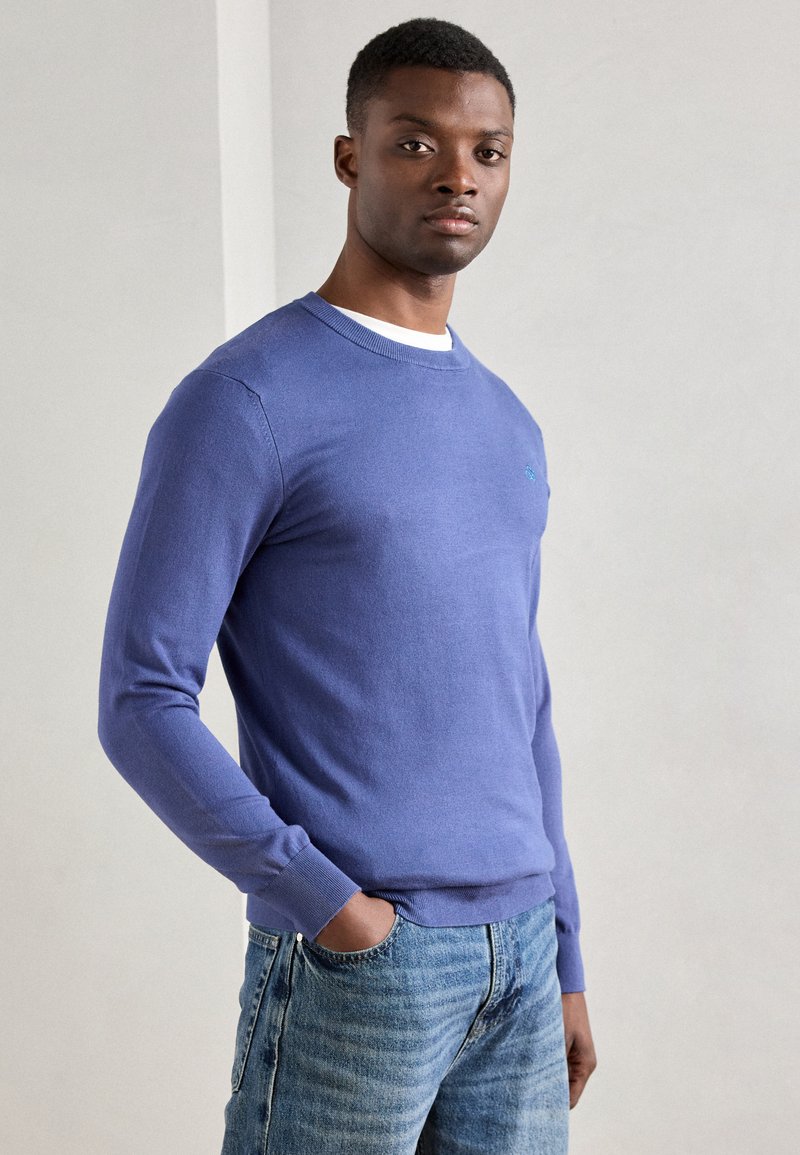 Hellblauer Strickpullover mit langen Ärmeln, rundem Ausschnitt und gerippten Bündchen. Verfügt über ein kleines Logo auf der Brust. Kombiniert mit hellblauen Jeans.