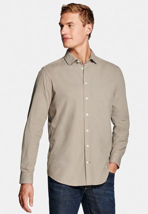 Man die een beige overhemd met lange mouwen en donkere jeans draagt, kijkt naar links met één hand in zijn zak.