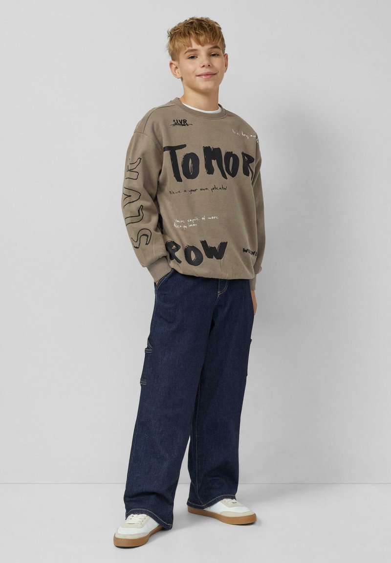 Bruine trui met gedurfde zwarte tekst, gecombineerd met losse donkerblauwe jeans en witte sneakers met beige accenten.