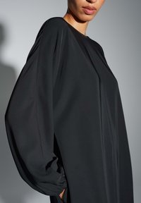Zwarte loszittende jurk met lange, volumineuze mouwen, gemaakt van gladde stof, met zijslits en gerimpelde mouwboorden.