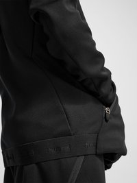 Person iført sort Hummel sportjakke med hånd i lynlåslomme i siden, vist fra taljen til midt på overkroppen mod hvid baggrund.