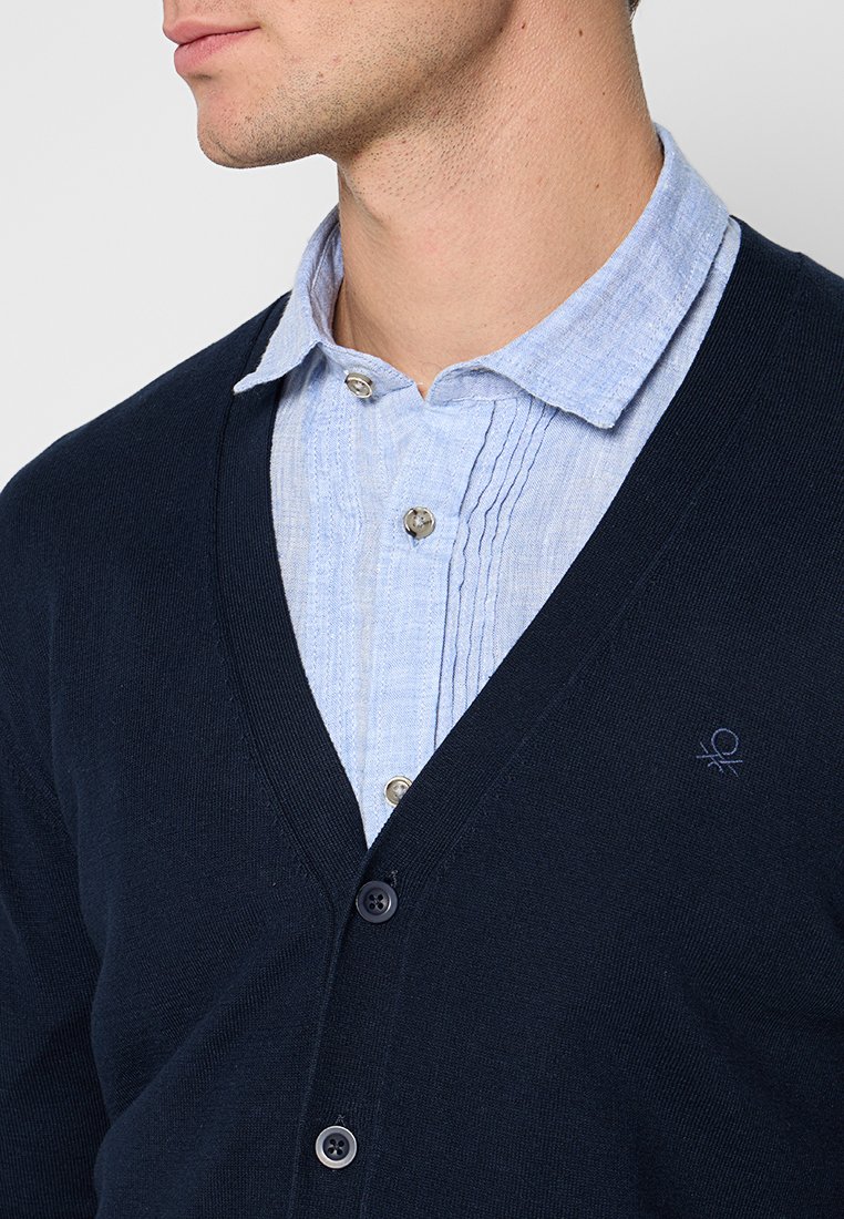 Cardigan lavorato a maglia blu navy con scollatura a V, dotato di bottoni, indossato sopra una camicia azzurra chiara con colletto, pieghe verticali e chiusura con bottoni.