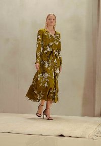 Vestido floral verde-oliva com mangas longas, decote em V e detalhe na cintura ajustada. Tecido suave e fluido com uma bainha de comprimento médio. Sapatos de salto pretos.