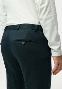 Dunkelgrüne Hose aus strukturiertem Stoff, mit zwei Gesäßtaschen mit Knopfverschluss und einer taillierten Passform.