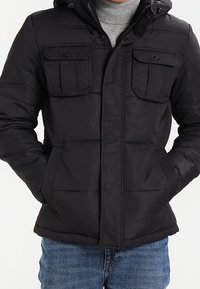 Svart vadderad jacka med huva, som har två bröstfickor, en framdragkedja och textilstruktur. Buren över en grå tröja och jeans.