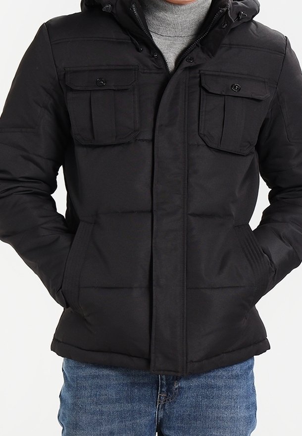 Svart vadderad jacka med huva, som har två bröstfickor, en framdragkedja och textilstruktur. Buren över en grå tröja och jeans.