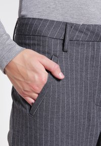 Grijze broek met krijtstrepen en een gladde textuur, met een zijzak en een op maat gemaakte pasvorm. Een hand rust in de zak, wat de ontwerpelementen benadrukt.