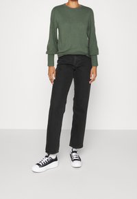 Pull en maille vert foncé avec des poignets côtelés, associé à un jean noir à jambes droites et des baskets noir et blanc. Texture lisse sur l'ensemble.