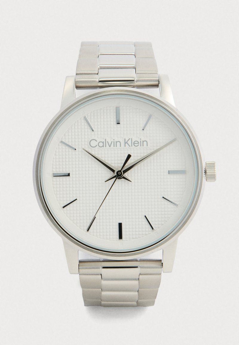 Montre-bracelet en métal argenté Calvin Klein avec un cadran blanc texturé, des index horaires minimalistes et trois aiguilles noires indiquant 10h10m35s.
