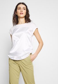 Top blanco de manga corta con un acabado satinado en la parte delantera y una textura mate en la parte trasera, combinado con pantalones de color caqui claro.