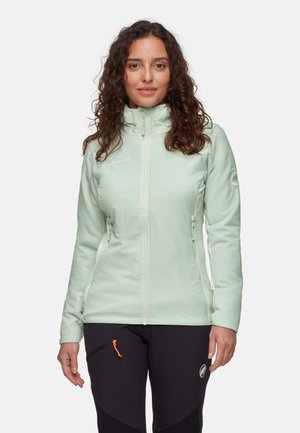 Vrouw met krullend haar, gekleed in een lichtgroene zip-up jas en zwarte outdoor broeken, tegen een effen witte achtergrond.