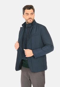 Giacca trapuntata blu navy con collo alto, chiusura a zip e tasche laterali. Tessuto liscio e design strutturato, adatta per un abbigliamento casual.