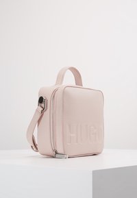Borsa quadrata compatta in pelle sintetica leggermente ruvida di colore rosa chiaro, dotata di manico superiore, zip laterale e logo "HUGO" impresso sul davanti.