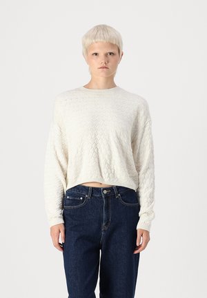 Hvit strikket genser med diamantmønster, rund halslinje og cropped hem. Kombinert med mørkeblå høyhårede jeans.