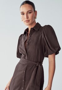 Robe marron à boutons avec des manches bouffantes, taille cintrée et ceinture à nouer. Fabriquée en tissu texturé, elle présente un décolleté plongeant et une longueur jusqu'aux genoux.