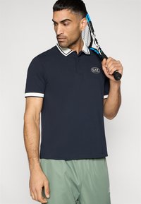 Ναυτικό μπλε πόλο μπλουζάκι με λευκές λεπτομέρειες και επωνυμία λογότυπου, κοντά μανίκια, σχέδιο με γιακά, φορεμένο με πράσινα αθλητικά σορτς, κρατώντας ρακέτα τένις.