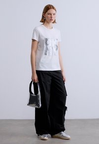 Λευκό βαμβακερό T-shirt με μεταλλικό λογότυπο "DKNY", σε συνδυασμό με φαρδιά μαύρα cargo παντελόνια. Το μοντέλο κρατάει μια μικρή, λαμπερή μαύρη τσάντα και φοράει ασημί αθλητικά παπούτσια.