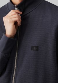 Mörk marinblå zip-up jacka med en strukturerad, våffelstickad design. Har en hög krage och en liten svart logotyp på bröstet.