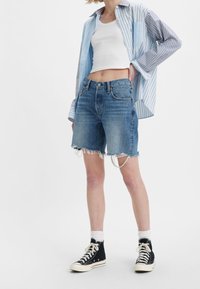 Shorts en denim avec des bords effilochés, délavés, et un design à cinq poches, associés à un crop top blanc et une chemise à rayures bleues à boutons.