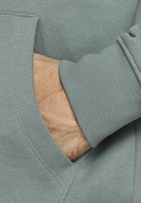 Zelená bavlněná mikina s vroubkovaným manžetem a přední kapsou, která ukazuje ruku odpočívající uvnitř kapsy s viditelnou texturou.