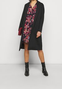 Schwarzer langer Mantel über einem bunten Blumenkleid, kombiniert mit schwarzen Stiefeletten. Das Outfit hat einen lässigen Schnitt und ein auffälliges, farbenfrohes Muster.
