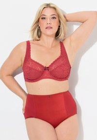 Reggiseno rosso in pizzo con bordi scallop e un fiocco decorativo al centro; abbinato a mutandine rosse a vita alta con pannelli laterali testurizzati.