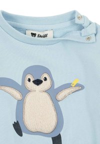 Camisa de manga corta azul claro con un aplique de pingüino en tela crema y gris, detalles bordados y una etiqueta amarilla cerca del cuello.