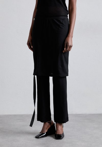 Ensemble noir composé d'un haut noir ajusté à col rond et d'une jupe noire courte superposée sur un pantalon large noir. Tissu texturé avec des détails à nouer.