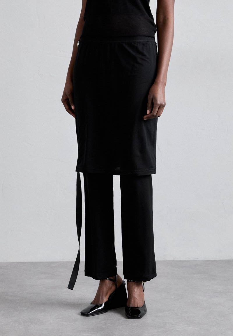 Conjunto preto com um top preto ajustado de gola redonda e uma saia preta curta sobre calças largas pretas. Tecido texturizado com detalhes em laço.