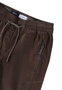 Pantalons marron avec taille élastique et cordon de serrage, dotés d'une poche zippée sur le côté et d'une texture lisse. Étiquette de marque visible à l'intérieur.