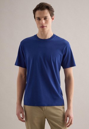 Joven con una camiseta azul lisa de manga corta y pantalones beige, de pie frente a un fondo liso de color claro.