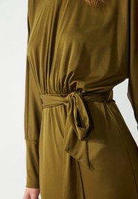 Olivengrønn kjole med lange ermer, som har et samlet bodice, et belte med knyting i midjen, og glatt, flytende stoff.