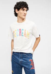 Λευκό β αμβακερό T-shirt με πολύχρωμο κείμενο "ICE CREAM"· συνδυασμένο με μπλε τζιν παντελόνι διακοσμημένο με καρικατούρες και γράμμα 'L'.