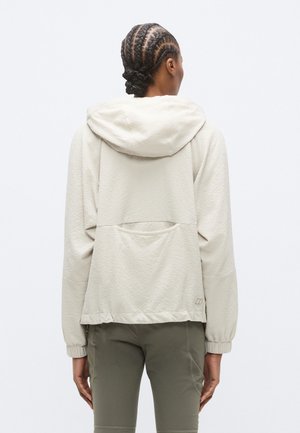 Veste à capuche beige clair en tissu texturé, avec poignets élastiques et détail de poche arrière. Capuche ajustable et coupe ample.
