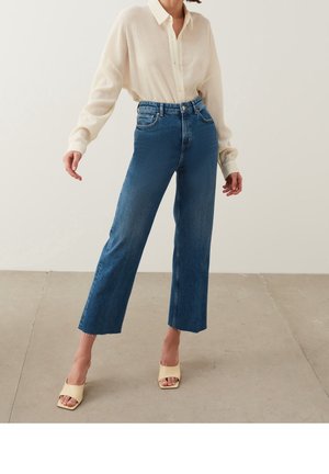 Nainen, joka pukeutunut kermanväriseen pitkähihaiseen teksturoituun puseroon, korkeavyötäröisiin lyhennettyihin sinisiin farkkuihin ja beigenvärisiin avokkaisiin sandaaleihin, seisoo laatoitetulla lattialla.