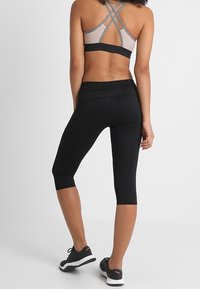 Nainen, jolla on päällään harmaa urheiluliivi, jossa on ristiin raskaavat hihat, ja mustat lyhyet leggingsit, seisoo tasaisella valkoisella taustalla.