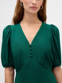 Woman wearing a green dress with puffed short sleeves, a V-neckline, and four fabric-covered buttons down the front.