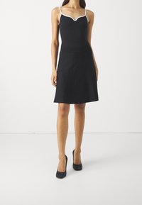 Schwarzes ärmelloses Kleid mit einem gewellten Ausschnitt, der mit weißem Saum verziert ist. Ein schwingender Rock endet auf Oberschenkelhöhe und wird mit schwarzen High Heels getragen.