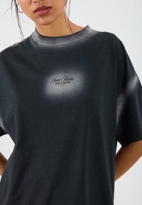 Sort oversized bomulds t-shirt med en falmet grå halsudskæring. Har et centreret logo i hvid tekst. Korte ærmer, glat tekstur.