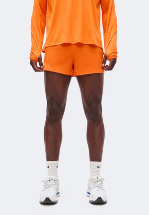 Mann iført en knalloransje atletisk langermet skjorte og shorts, hvite Nike-sokker og hvite løpetreningssko, stående foran ensfarget bakgrunn.