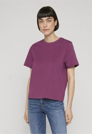 Mujer con cabello corto y oscuro que lleva una camiseta morada lisa de manga corta y vaqueros azules, de pie frente a un fondo gris claro.