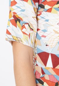 Camisa de manga corta con un colorido patrón geométrico que presenta formas rojas, amarillas, azules y negras sobre un fondo blanco. Textura de tela ligera.