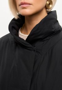 Zbliżenie osoby noszącej czarną kurtkę z wysokim kołnierzem, duży okrągły złoty kolczyk oraz złoty naszyjnik na białym tle.