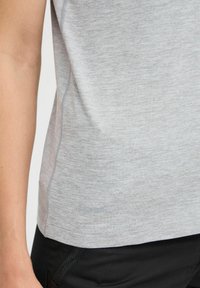 Maglietta sportiva grigio chiaro in tessuto testurizzato, con cuciture sottili e orlo curvo, indossata con pantaloni neri.