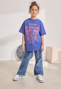 Modra grafična majica s kratkimi rokavi in napisom "Foo Fighters", nošena z raztrganimi jeansa in belimi supergami. Ima kratke rokave in sproščen kroj.